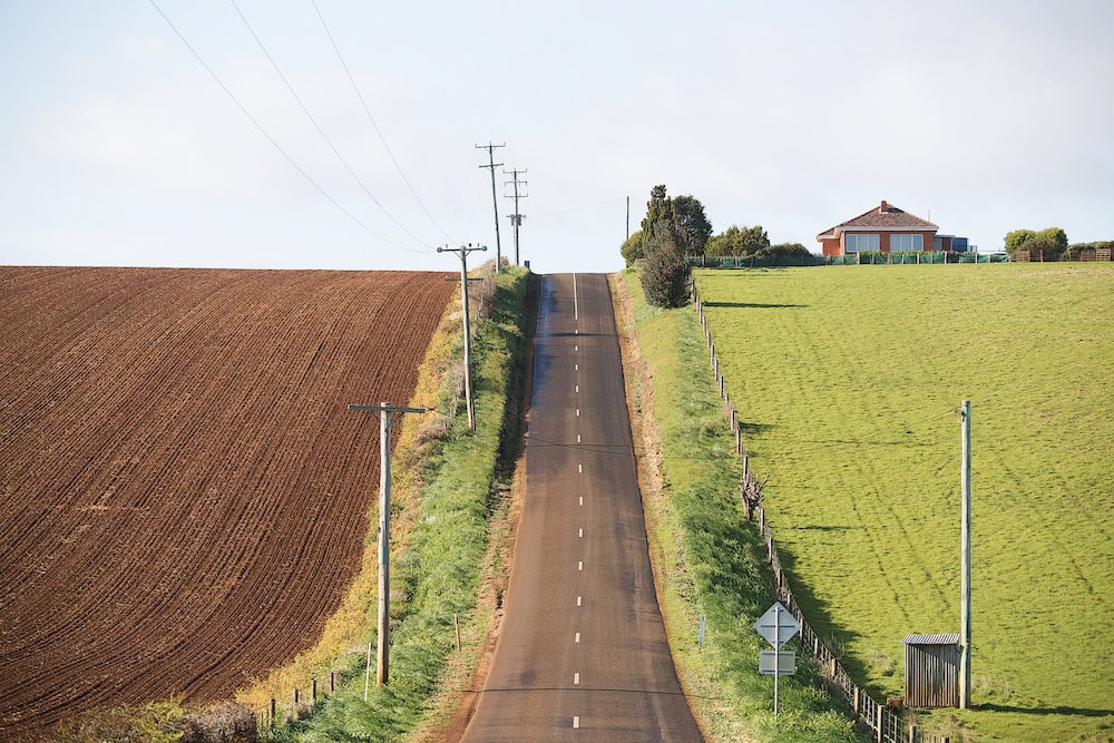 Rural road past ploughed field on one side and green pasture on the other.